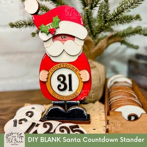 May include: A DIY Santa countdown stander with a red and white Santa Claus design. The stander features a countdown dial with the number 31, and includes numbered wooden discs. The text "COUNTDOWN TO CHRISTMAS" is visible.
