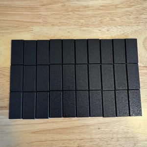 May include: Twenty-one black rectangular game pieces arranged in three rows on a light brown wooden surface. The pieces have a textured surface and are evenly spaced, suggesting a board game or puzzle.