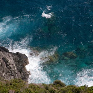 May include: A rocky cliff face with dark grey and brown rock formations. The cliff face is partially submerged in a body of water with blue and white waves crashing against the rocks.