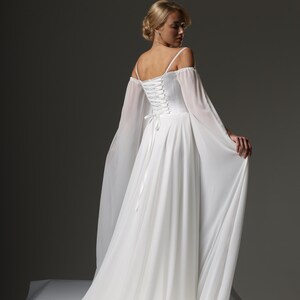 May include: A white wedding dress with a lace-up back and off-the-shoulder sleeves. The dress has a flowing skirt and a long train.