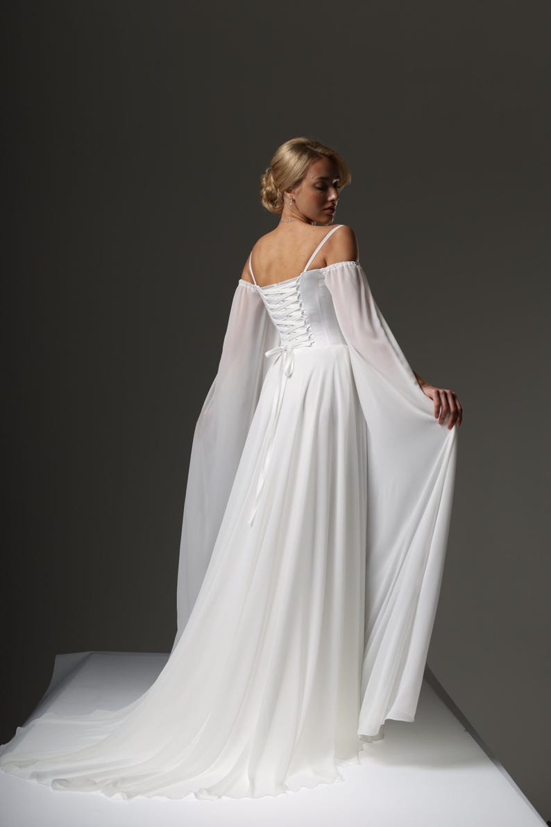 May include: A white wedding dress with a lace-up back and off-the-shoulder sleeves. The dress has a flowing skirt and a long train.