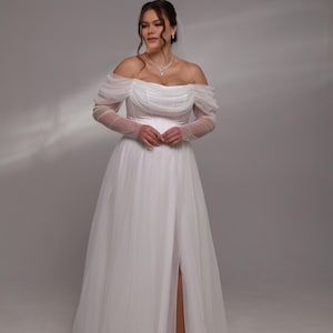 May include: A white off-the-shoulder wedding dress with long sheer sleeves and a slit in the skirt.
