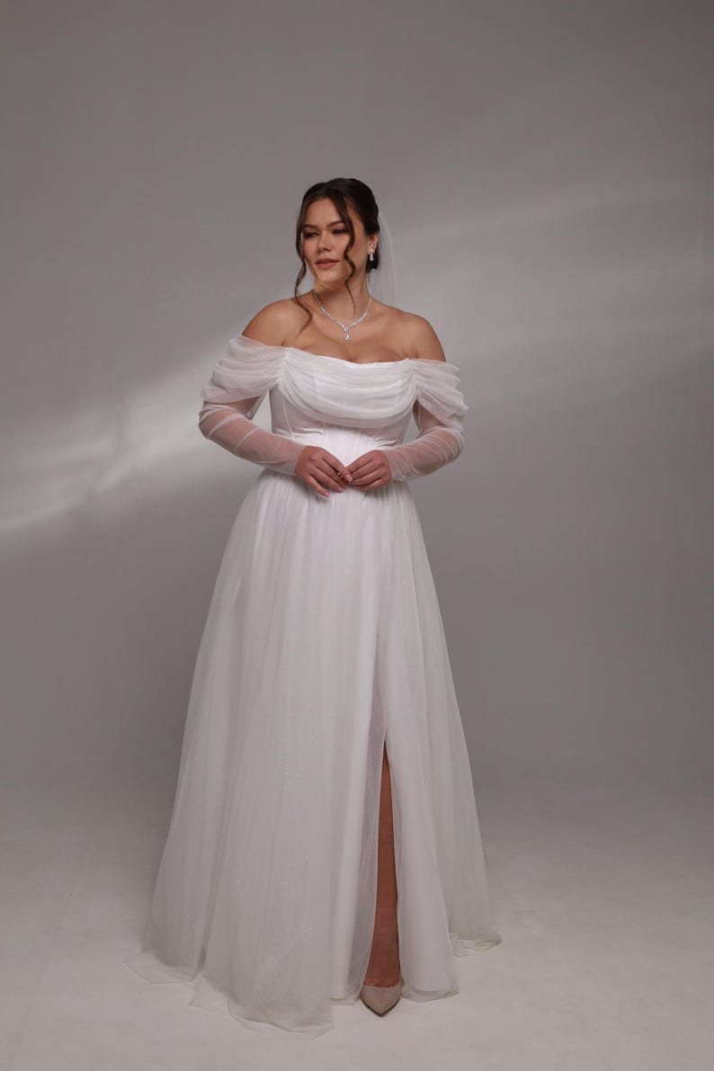 May include: A white off-the-shoulder wedding dress with long sheer sleeves and a slit in the skirt.