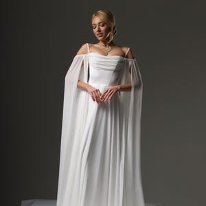 May include: A white wedding dress with a flowing chiffon skirt and off-the-shoulder straps. The dress has a fitted bodice and a delicate, flowing cape.