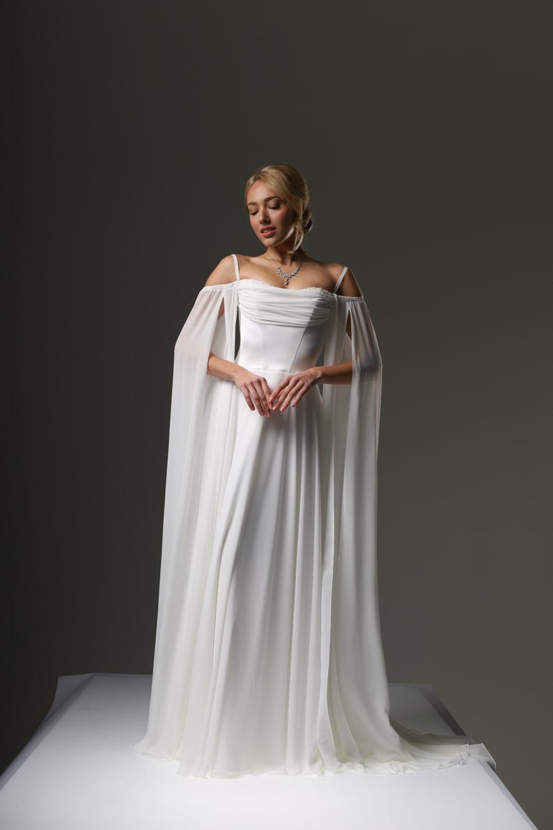 May include: A white wedding dress with a flowing chiffon skirt and off-the-shoulder straps. The dress has a fitted bodice and a delicate, flowing cape.