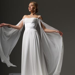 May include: A white wedding dress with a fitted bodice and a flowing skirt. The dress has off-the-shoulder straps and a long, flowing train.