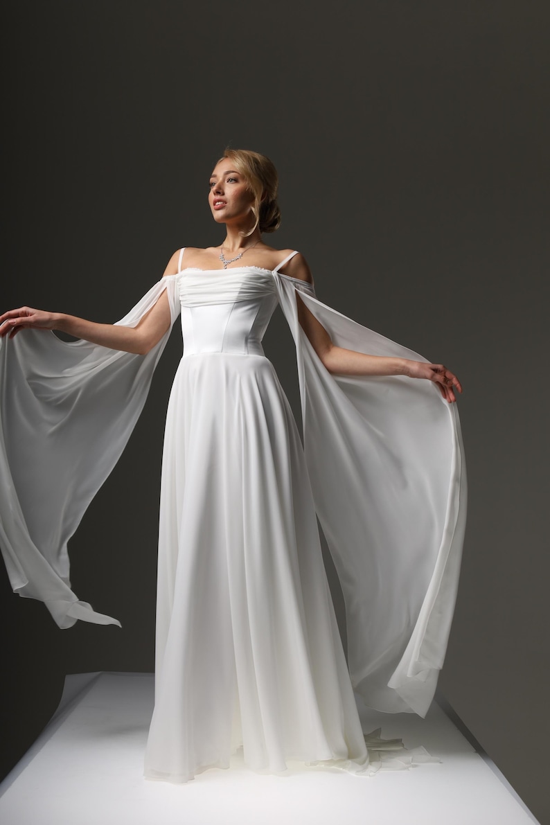 May include: A white wedding dress with a fitted bodice and a flowing skirt. The dress has off-the-shoulder straps and a long, flowing train.