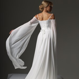 May include: A white wedding dress with a lace-up back and long, flowing sleeves. The dress has a simple, elegant design and is perfect for a beach wedding or a casual ceremony.