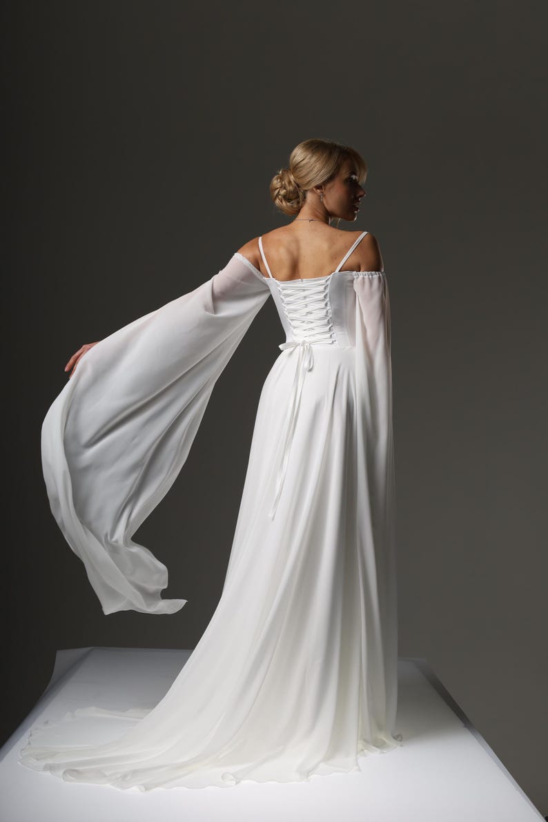 May include: A white wedding dress with a lace-up back and long, flowing sleeves. The dress has a simple, elegant design and is perfect for a beach wedding or a casual ceremony.