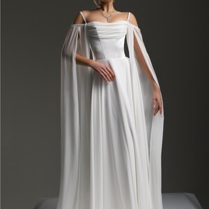 May include: A white wedding dress with a flowing skirt and off-the-shoulder straps. The dress has a fitted bodice and a long, flowing train.