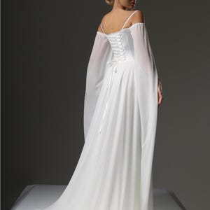May include: A white off-the-shoulder wedding dress with a lace-up back and long flowing sleeves. The dress has a flowing skirt and a fitted bodice.