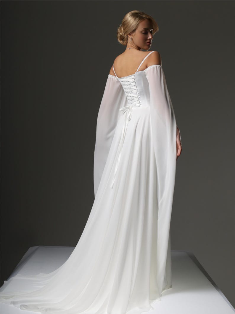 May include: A white off-the-shoulder wedding dress with a lace-up back and long flowing sleeves. The dress has a flowing skirt and a fitted bodice.