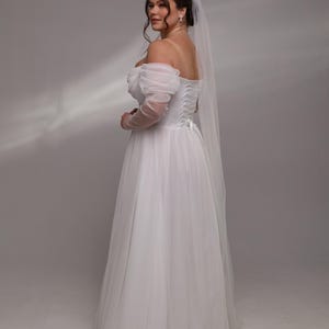 May include: A white off-the-shoulder wedding dress with a tulle skirt and a lace-up back. The dress has a veil attached to the back of the head.