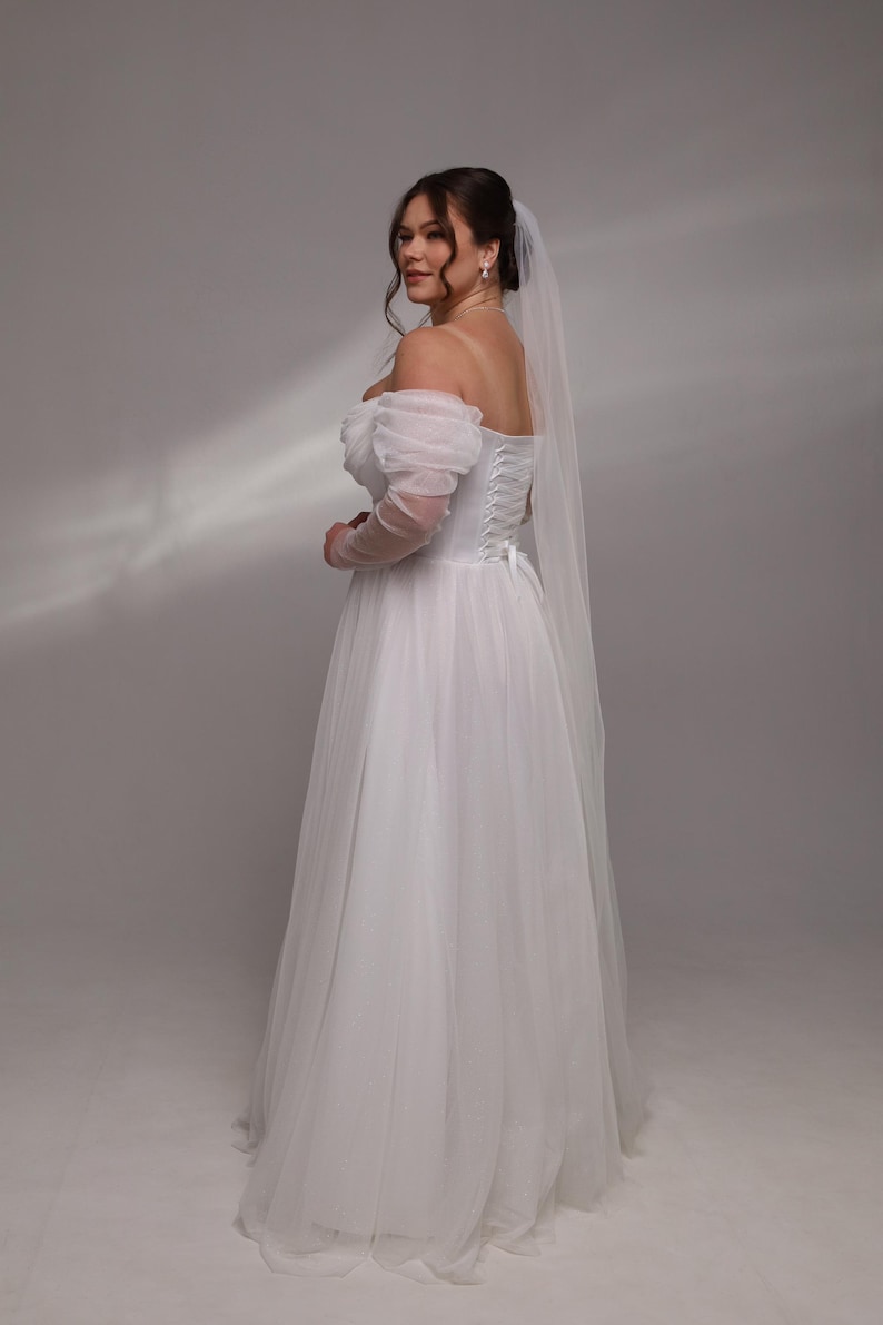 May include: A white off-the-shoulder wedding dress with a tulle skirt and a lace-up back. The dress has a veil attached to the back of the head.