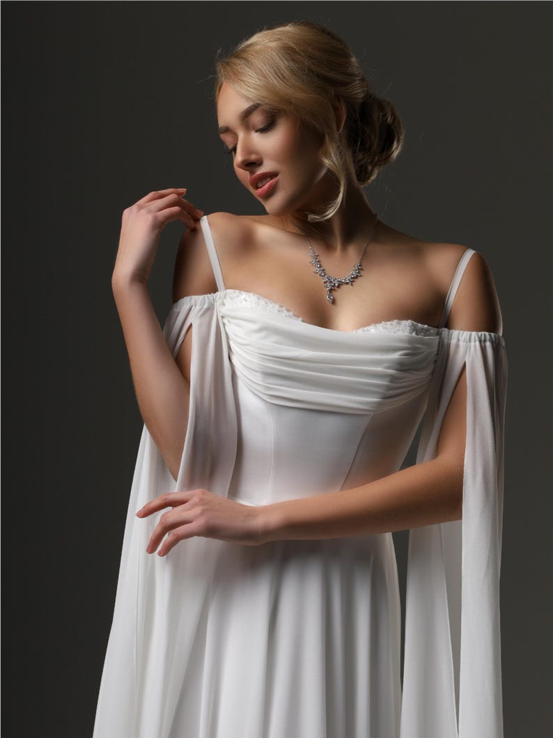 May include: A white wedding dress with a sweetheart neckline and a flowing skirt. The dress has sheer, flowing sleeves that drape over the shoulders. The model is wearing a delicate silver necklace.
