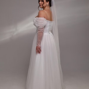 May include: A white tulle wedding dress with a sweetheart neckline and puff sleeves. The dress has a corset back and a long train. The bride is wearing a white veil.