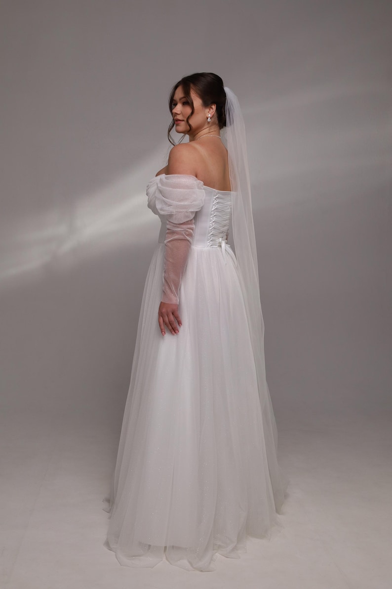 May include: A white tulle wedding dress with a sweetheart neckline and puff sleeves. The dress has a corset back and a long train. The bride is wearing a white veil.