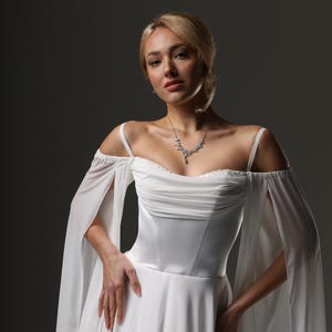 May include: A woman wearing a white wedding dress with a sweetheart neckline and sheer, off-the-shoulder sleeves. The dress has a fitted bodice and a flowing skirt. She is wearing a delicate silver necklace.