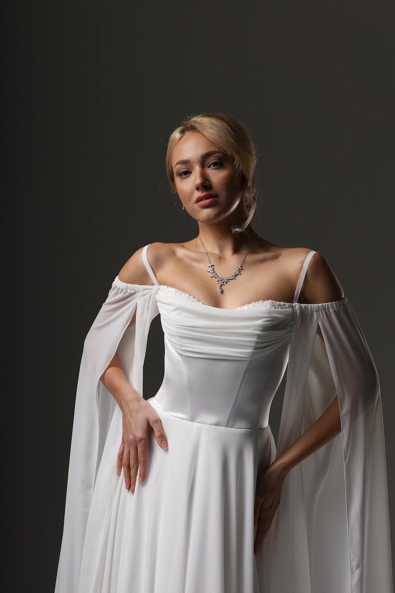 May include: A woman wearing a white wedding dress with a sweetheart neckline and sheer, off-the-shoulder sleeves. The dress has a fitted bodice and a flowing skirt. She is wearing a delicate silver necklace.