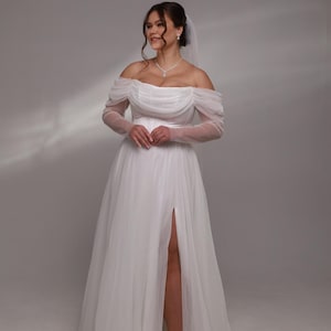 May include: A white off-the-shoulder wedding dress with long sheer sleeves and a thigh-high slit. The dress has a flowing skirt and is made of a soft, delicate fabric.