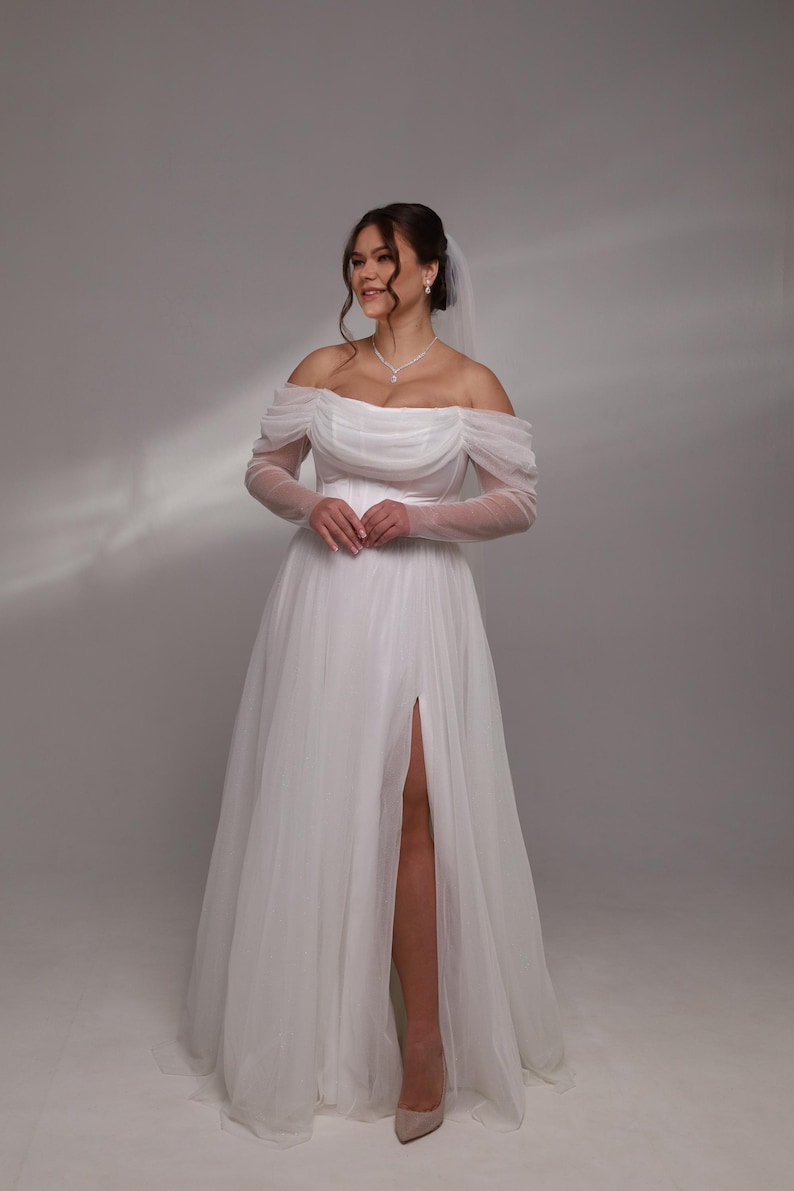 May include: A white off-the-shoulder wedding dress with long sheer sleeves and a thigh-high slit. The dress has a flowing skirt and is made of a soft, delicate fabric.