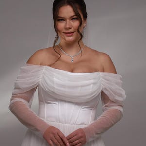 May include: A white off-the-shoulder wedding dress with tulle sleeves and a fitted bodice. The dress has a flowing skirt and is decorated with small, sparkling details.