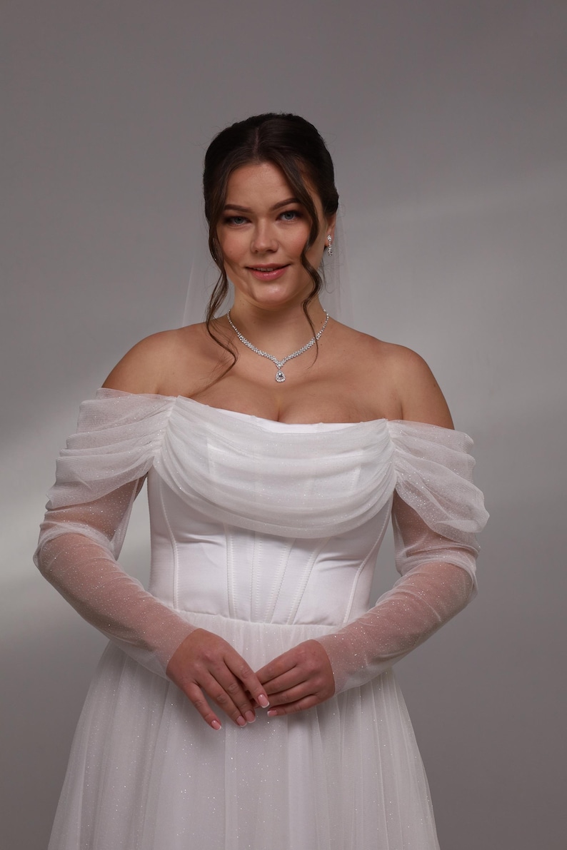 May include: A white off-the-shoulder wedding dress with tulle sleeves and a fitted bodice. The dress has a flowing skirt and is decorated with small, sparkling details.