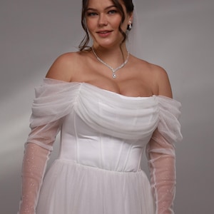 May include: A white off-the-shoulder wedding dress with a tulle skirt and long sleeves. The sleeves are made of a sheer fabric with small white dots. The dress has a fitted bodice with a sweetheart neckline and a corset-style closure.