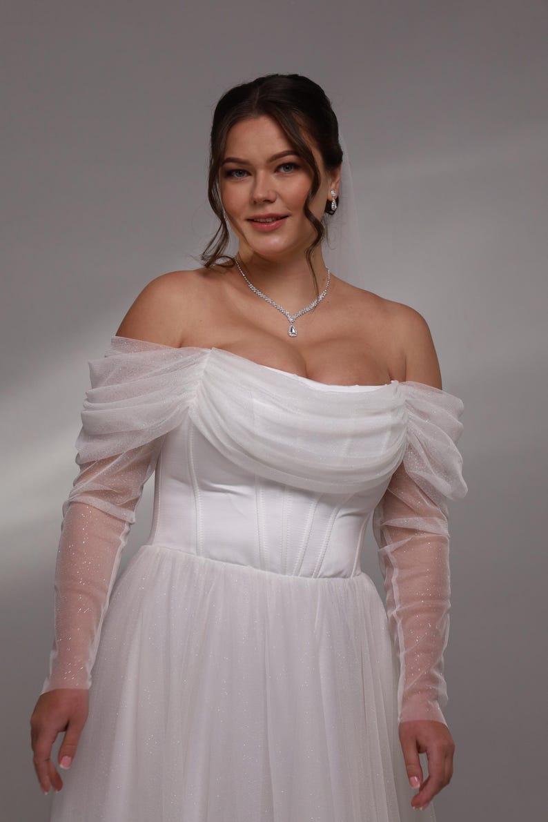 May include: A white off-the-shoulder wedding dress with a tulle skirt and long sleeves. The sleeves are made of a sheer fabric with small white dots. The dress has a fitted bodice with a sweetheart neckline and a corset-style closure.
