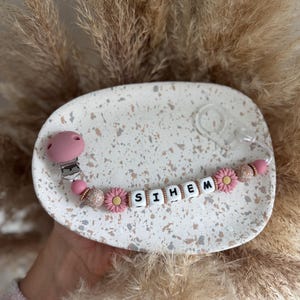 Op de afbeelding: Een roze en witte fopspeenclip voor baby's met de naam "SIHEM" gespeld in witte letterkralen. De clip heeft roze madeliefvormige kralen en een roze fopspeenhouder. De clip ligt op een gespikkelde witte plaat.