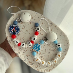Op de afbeelding: Twee baby fopspeenclips met witte, blauwe, rode en zwarte kralen. Eén clip heeft een Paris Saint-Germain logo, de andere een Olympique de Marseille logo. De naam "JULES" staat op de ene clip, en "NUZZ" op de andere.