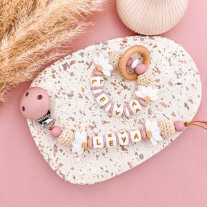 Peut inclure: Une attache-tétine et un anneau de dentition assorti avec des fleurs marguerites en silicone blanc, des perles en silicone rose poudré, des perles crochetées beiges et des cubes blancs avec le nom LEYA inscrit en lettres dorées.