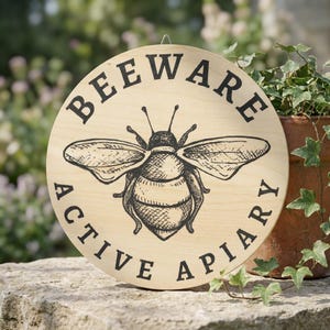 Può includere: Cartello rotondo in legno con la scritta "BEEWARE ACTIVE APIARY" in nero. Un'illustrazione dettagliata di un'ape è al centro. Il cartello è appeso all'aperto vicino a una pianta in vaso.