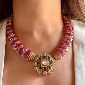 May include: A luxurious necklace featuring a central floral pendant with a large, clear center stone surrounded by smaller green stones. The necklace's curved sections are adorned with numerous pink gemstones, creating a bold and elegant statement piece.