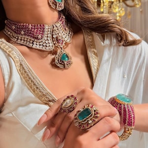 May include: A luxurious jewellery set comprising a choker necklace, earrings, rings, and a bracelet. The set features emerald green and ruby red gemstones, set in gold. The necklace and bracelet have a wide band design, and the rings are large and striking.