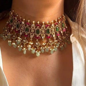 Op de afbeelding: Een goudkleurige choker ketting met smaragdgroene en robijnrode accenten. De ketting heeft vierkante en druppelvormige edelstenen, met bungelende groene kralen. Het ontwerp is ingewikkeld en heeft een vintage uitstraling.