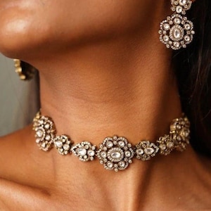 May include: A gold choker necklace with a delicate design featuring multiple white gemstones. The necklace is made of metal and has a floral pattern.
