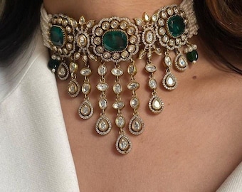 May include: A gold choker necklace with a white pearl base and emerald green stones. The necklace has a dangling design with smaller clear stones.