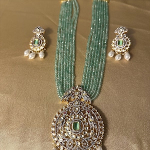 May include: A multi-strand necklace with a large, ornate pendant featuring a green gemstone surrounded by smaller stones. The pendant is attached to a chain of green beads and has a pearl drop. The necklace is accompanied by matching earrings with green gemstones and pearl drops.