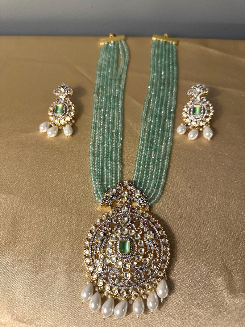 May include: A multi-strand necklace with a large, ornate pendant featuring a green gemstone surrounded by smaller stones. The pendant is attached to a chain of green beads and has a pearl drop. The necklace is accompanied by matching earrings with green gemstones and pearl drops.