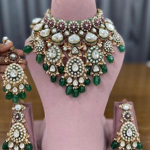 Puede incluir: Un collar dorado y un juego de pendientes a juego. La joyer&iacute;a presenta piedras grandes y claras en forma de l&aacute;grima, rodeadas de piedras blancas m&aacute;s peque&ntilde;as y detalles verdes. El conjunto se exhibe en un maniqu&iacute; rosa.