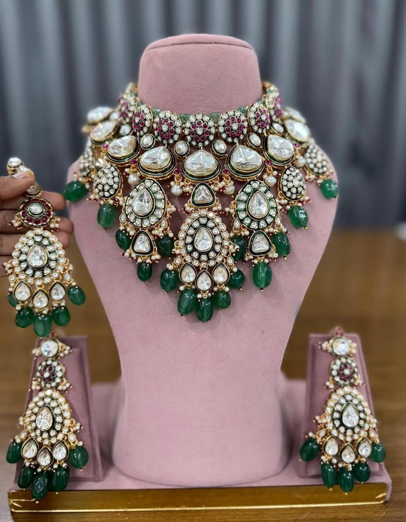 Puede incluir: Un collar dorado y un juego de pendientes a juego. La joyer&iacute;a presenta piedras grandes y claras en forma de l&aacute;grima, rodeadas de piedras blancas m&aacute;s peque&ntilde;as y detalles verdes. El conjunto se exhibe en un maniqu&iacute; rosa.