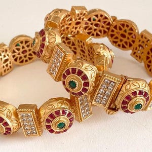 May include: Two gold bangles with intricate designs, featuring red and green gemstones. The bangles are adorned with filigree work and have a traditional Indian style.