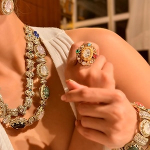 Puede incluir: Un anillo dorado adornado con una gran piedra central transparente, rodeada de piedras preciosas de colores más pequeñas. También se ven un brazalete y un collar a juego, con diseños y piedras similares. La joyería se encuentra sobre un fondo suave y neutro.