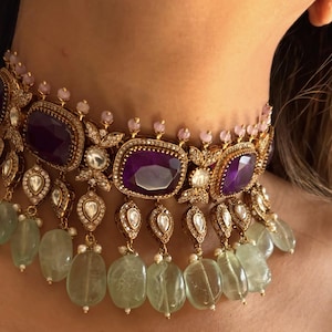 May include: An ornate gold choker necklace with large, rectangular, deep purple gemstones. The necklace features smaller clear and pink stones, and dangling green-tinted teardrop-shaped beads. The design is intricate and detailed.