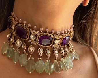 May include: An ornate gold choker necklace with large, rectangular, deep purple gemstones. The necklace features smaller clear and pink stones, and dangling green-tinted teardrop-shaped beads. The design is intricate and detailed.