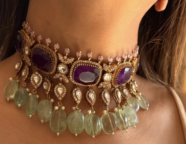 Puede incluir: Un collar gargantilla dorado con grandes piedras preciosas rectangulares de color p&uacute;rpura intenso. El collar est&aacute; adornado con piedras m&aacute;s peque&ntilde;as transparentes y rosas, y cuentas colgantes en forma de l&aacute;grima de color verde. El dise&ntilde;o es intrincado y ornamentado.