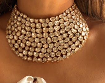 May include: A wide, gold-toned choker necklace densely set with numerous clear, irregularly shaped gemstones. The necklace has a curved shape, designed to sit around the base of the neck. The gemstones are closely packed together, creating a sparkling effect.