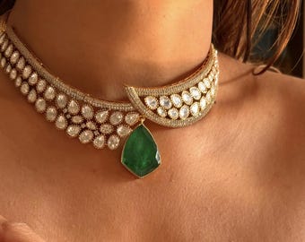 May include: A gold-toned statement necklace featuring a series of clear, teardrop-shaped stones and smaller, round stones. A large, green, pear-shaped pendant hangs from the center. The necklace is worn around the neck.