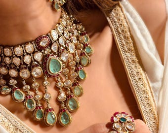 May include: A gold-toned necklace and earring set featuring uncut stones in shades of mint green and deep red. The necklace is a multi-layered choker design with dangling teardrop-shaped pendants, while the ring features a bird-shaped motif.
